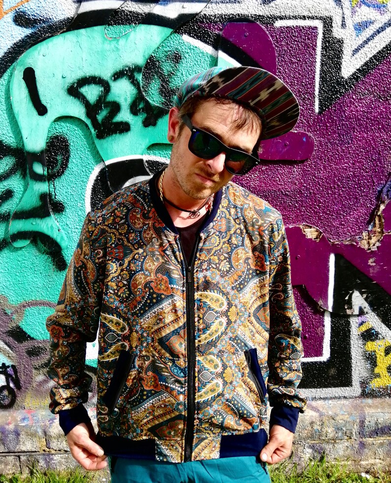 May include: A man wearing a paisley patterned bomber jacket with a zipper closure. The jacket has a blue, gold, and brown color scheme. He is wearing a blue and black patterned hat and sunglasses.