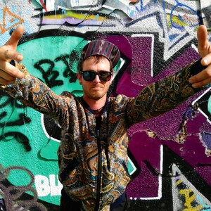 May include: A man wearing a patterned jacket, sunglasses, and a hat with a paisley design. He is standing in front of a graffiti wall with his arms outstretched.