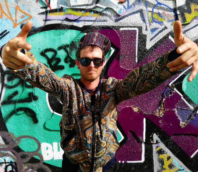 May include: A man wearing a patterned jacket, sunglasses, and a hat with a paisley design. He is standing in front of a graffiti wall with his arms outstretched.