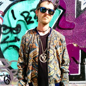 May include: A man wearing a patterned bomber jacket with a gold chain necklace and a large pendant. He is wearing a black t-shirt, blue jeans, and sunglasses. He is standing in front of a graffiti wall.