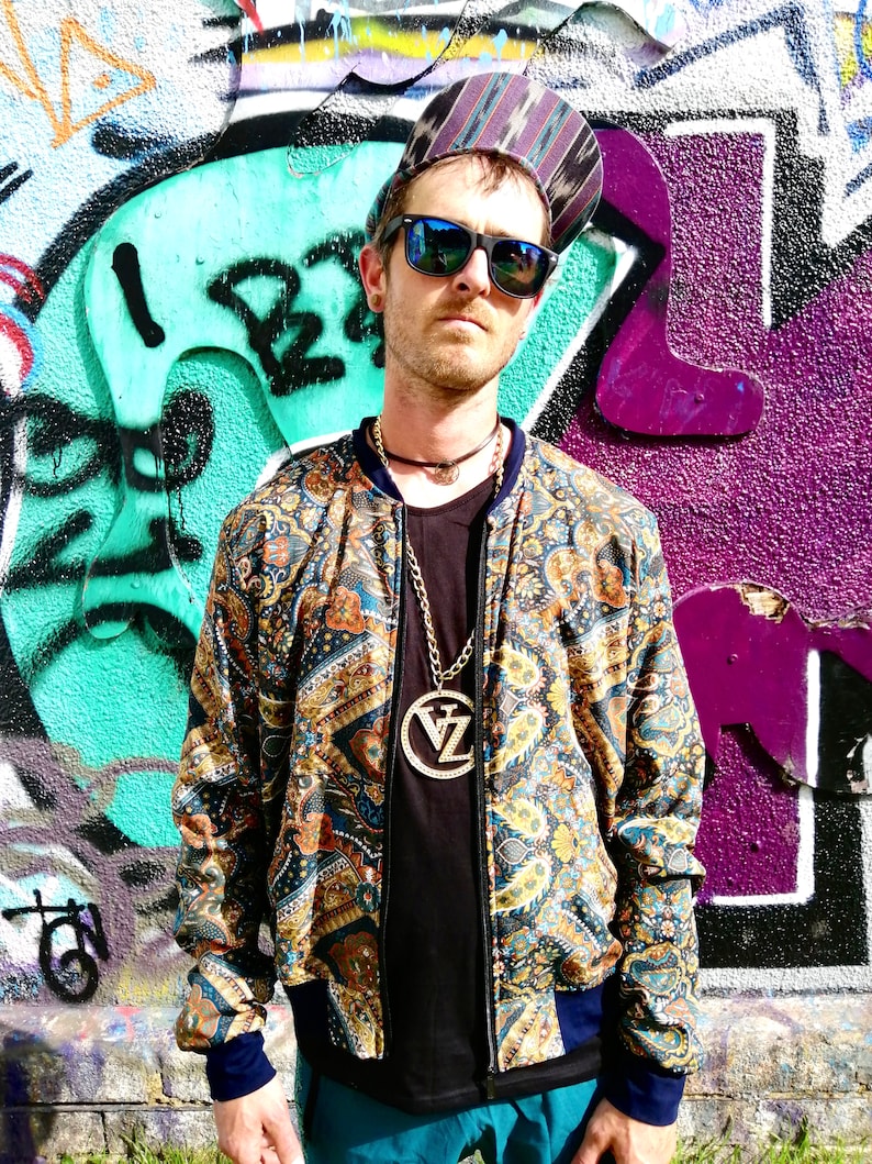 May include: A man wearing a patterned bomber jacket with a gold chain necklace and a large pendant. He is wearing a black t-shirt, blue jeans, and sunglasses. He is standing in front of a graffiti wall.