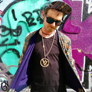 May include: A person wearing a black shirt with a gold chain necklace and a large gold pendant with the letters "VZ" inside. The person is wearing a patterned jacket with the lining showing. The person is wearing a blue and green patterned hat.