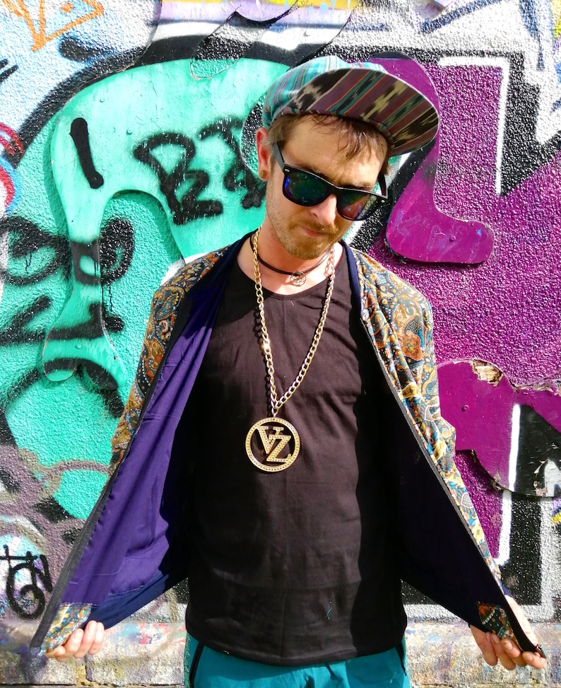 May include: A person wearing a black shirt with a gold chain necklace and a large gold pendant with the letters "VZ" inside. The person is wearing a patterned jacket with the lining showing. The person is wearing a blue and green patterned hat.