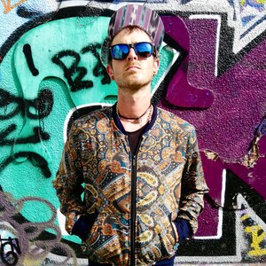 May include: A man wearing a blue, brown, and gold paisley patterned bomber jacket with a zipper closure. He is wearing a blue and black patterned hat and blue sunglasses. The man is standing in front of a graffiti wall.