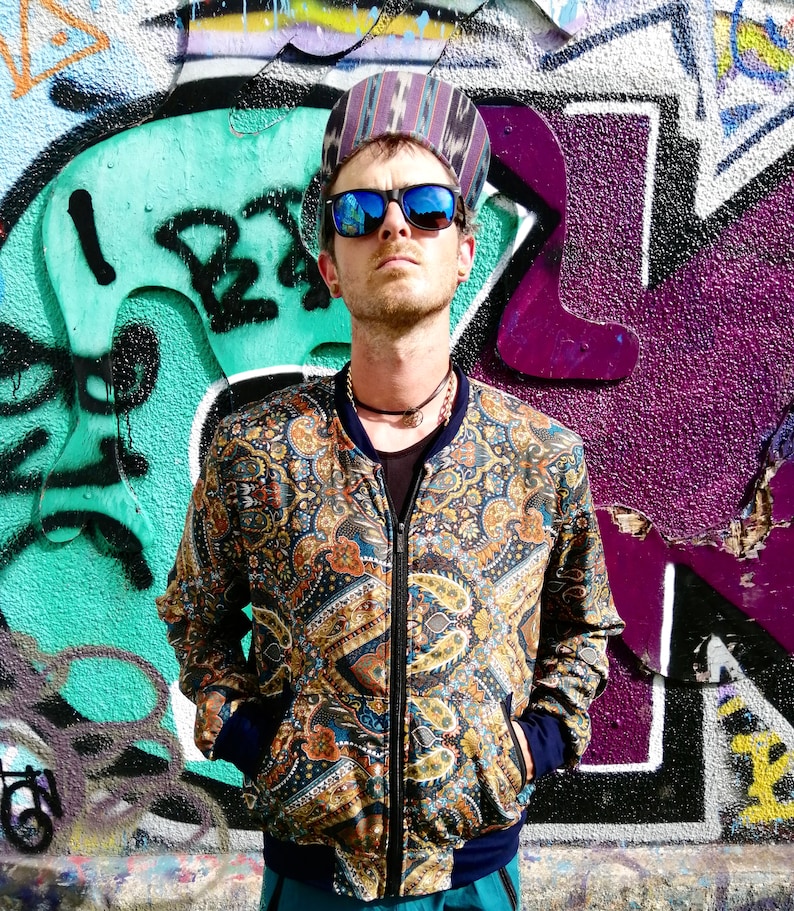May include: A man wearing a blue, brown, and gold paisley patterned bomber jacket with a zipper closure. He is wearing a blue and black patterned hat and blue sunglasses. The man is standing in front of a graffiti wall.
