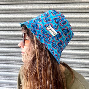 Könnte beinhalten: Ein blauer und roter Bucket-Hut mit geometrischem Muster. Der Hut hat ein kleines weißes Etikett mit dem Wort "Jackaranda" darauf.