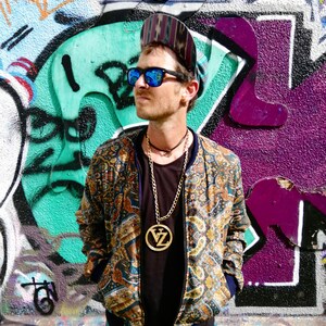 May include: A person wearing a patterned bomber jacket with a gold chain necklace and a large pendant. The pendant has the letters "VZ" on it. The person is wearing a blue and black hat and blue sunglasses.