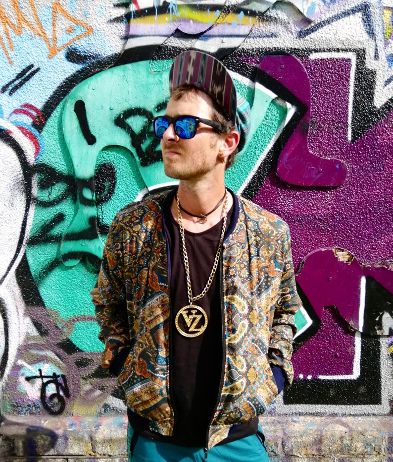 May include: A person wearing a patterned bomber jacket with a gold chain necklace and a large pendant. The pendant has the letters "VZ" on it. The person is wearing a blue and black hat and blue sunglasses.