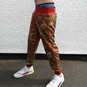 Op de afbeelding: Een rode en bruine broek met paisley print en een rode tailleband. De broek wordt gedragen door een persoon en de afbeelding is geknipt bij de enkels.