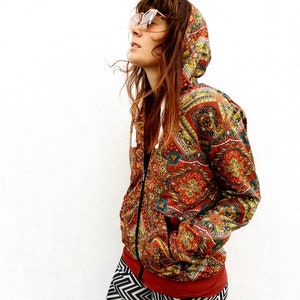 Puede incluir: Chaqueta con capucha y cremallera con estampado de cachemira en tonos rojos, dorados y azules. La chaqueta tiene una capucha con cordón y un ribete rojo oscuro. La persona lleva leggings estampados en blanco y negro y gafas de sol.
