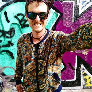 May include: A person wearing a paisley patterned bomber jacket with a zipper closure. The jacket has a blue collar and a blue waistband. The person is also wearing a black and white striped hat and sunglasses.