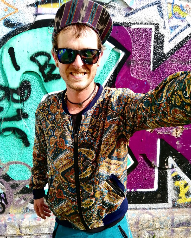 May include: A person wearing a paisley patterned bomber jacket with a zipper closure. The jacket has a blue collar and a blue waistband. The person is also wearing a black and white striped hat and sunglasses.