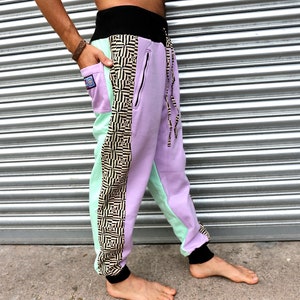 May include: Purple and mint green sweatpants with a black and white geometric pattern on the side. The sweatpants have a black waistband and a drawstring.