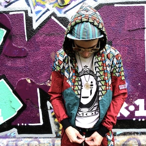 May include: A person wearing a colourful hooded jacket with a geometric pattern. The jacket has a red, green and blue colour scheme. The person is wearing a white t-shirt with the text "Est. 2011" and "MOR" printed on it. The person is standing in front of a graffiti wall.