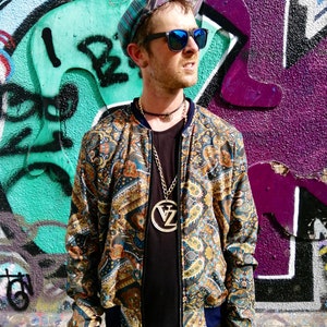 May include: A man wearing a patterned bomber jacket with a gold chain necklace and a large pendant. He is wearing a black t-shirt, teal pants, and a patterned hat. He is standing in front of a graffiti wall.