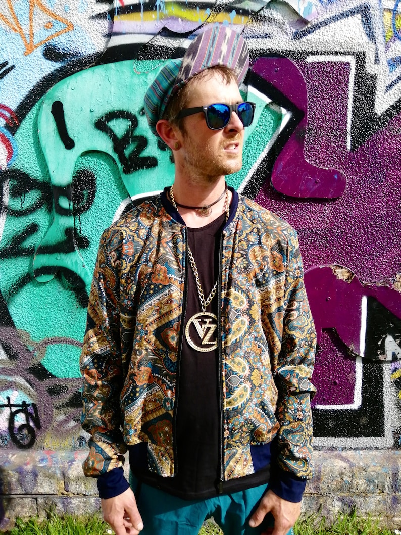 May include: A man wearing a patterned bomber jacket with a gold chain necklace and a large pendant. He is wearing a black t-shirt, teal pants, and a patterned hat. He is standing in front of a graffiti wall.