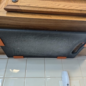 May include: A black cutting board with orange clips is attached to the underside of a wooden cabinet. The cutting board is hanging over a white tiled backsplash.