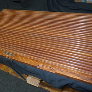 May include: A vintage wooden roll-top desk cover with a warm brown finish. The cover features a series of parallel, grooved slats. A small, ornate metal clasp is visible on the lower edge. The desk cover is designed to slide open and closed.