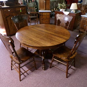 May include: A round wooden dining table with a central pedestal base featuring carved claw feet, accompanied by four matching wooden chairs. Each chair has a high backrest with intricate carved detailing and a contoured wooden seat.