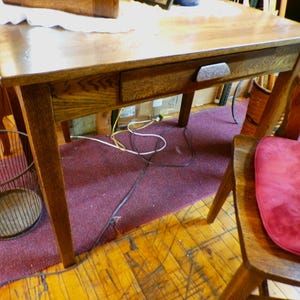 May include: A wooden desk with a single drawer and a metal drawer pull. The desk has a dark wood finish and is sitting on a red rug. There are electrical cords visible under the desk.