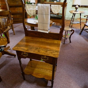 May include: An antique wooden washstand with a towel rack, drawer, and lower shelf. The washstand has a warm, medium-brown finish with visible wood grain. The drawer features decorative hardware. A checkered towel hangs from the rack. The washstand is a vintage furniture piece.