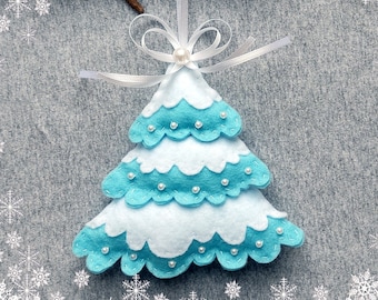 May include: A blue and white felt Christmas tree ornament with a white ribbon and pearl bead. The ornament is hanging from a branch against a gray background with a white snowflake border.