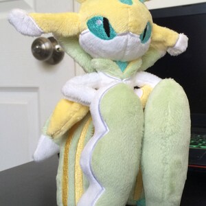 May include: A plush toy of a light green and yellow Pokemon with a white face and blue eyes. The toy has a long, flowing tail and is standing on a black surface.