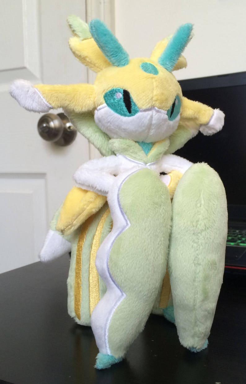 May include: A plush toy of a light green and yellow Pokemon with a white face and blue eyes. The toy has a long, flowing tail and is standing on a black surface.