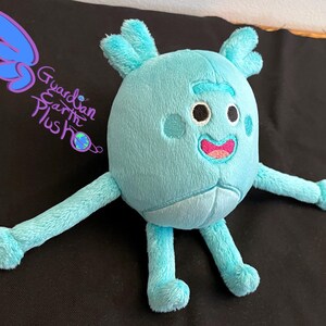Puede incluir: Un juguete de peluche azul claro con una cara sonriente, dos brazos y dos piernas. El juguete tiene un cuerpo redondo y una cabeza ligeramente puntiaguda. El juguete est&aacute; sentado sobre una superficie negra.