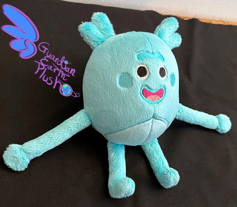 Puede incluir: Un juguete de peluche azul claro con una cara sonriente, dos brazos y dos piernas. El juguete tiene un cuerpo redondo y una cabeza ligeramente puntiaguda. El juguete est&aacute; sentado sobre una superficie negra.