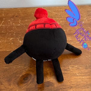 Puede incluir: Juguete de peluche negro con un sombrero rojo y blanco. El juguete tiene dos brazos negros y dos piernas negras. El juguete est&aacute; sentado sobre una superficie de madera.