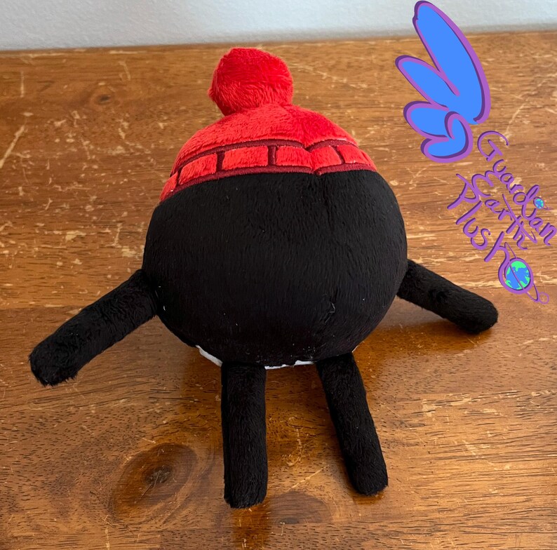 Puede incluir: Juguete de peluche negro con un sombrero rojo y blanco. El juguete tiene dos brazos negros y dos piernas negras. El juguete est&aacute; sentado sobre una superficie de madera.