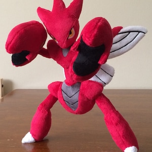 K&ouml;nnte beinhalten: Rotes und schwarzes Pl&uuml;schtier eines Pokemon-Charakters mit gro&szlig;en Armen und einem Fl&uuml;gel auf dem R&uuml;cken. Das Spielzeug steht auf einer Holzoberfl&auml;che.