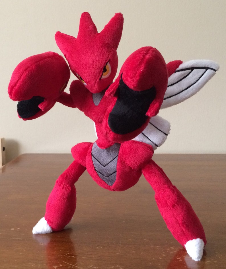 K&ouml;nnte beinhalten: Rotes und schwarzes Pl&uuml;schtier eines Pokemon-Charakters mit gro&szlig;en Armen und einem Fl&uuml;gel auf dem R&uuml;cken. Das Spielzeug steht auf einer Holzoberfl&auml;che.