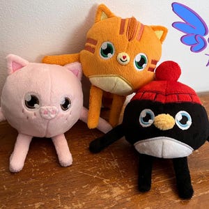 Puede incluir: Tres juguetes de peluche: un cerdo rosa, un gato naranja con rayas y un ping&uuml;ino negro con un gorro rojo. Todos los juguetes sonr&iacute;en y tienen ojos grandes. Los juguetes est&aacute;n sentados sobre una superficie de madera.