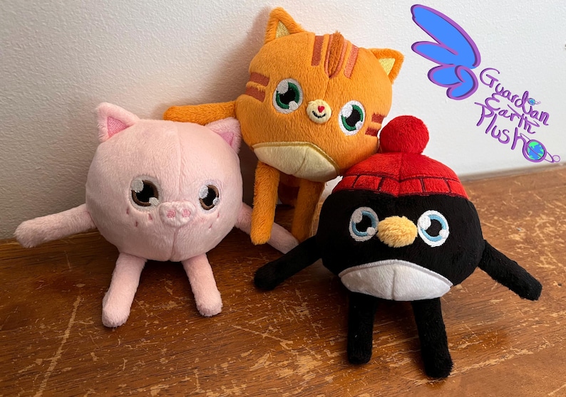 Puede incluir: Tres juguetes de peluche: un cerdo rosa, un gato naranja con rayas y un ping&uuml;ino negro con un gorro rojo. Todos los juguetes sonr&iacute;en y tienen ojos grandes. Los juguetes est&aacute;n sentados sobre una superficie de madera.