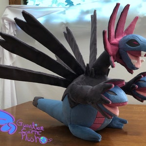 May include: A plush toy of the Pokemon Hydreigon. The toy is blue, black, and pink with three heads. The toy is posed with its wings spread out.