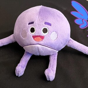 Puede incluir: Juguete de peluche morado con un cuerpo redondo y dos brazos largos. El juguete tiene una cara sonriente con dos ojos, una nariz y una boca. El juguete est&aacute; etiquetado como "Guardian Earth Plush" en el lado derecho.