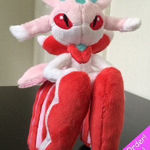 May include: Pink and red plush toy with a teal and white design. The toy has large red eyes and a teal and white crown. The toy is sitting on a black surface. Made to Order! 2-3 weeks!