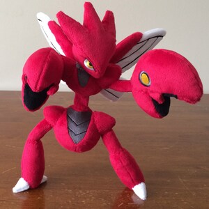 K&ouml;nnte beinhalten: Rotes und schwarzes Pl&uuml;schtier eines Pokemon-Charakters mit gro&szlig;en Krallen und Fl&uuml;geln. Das Spielzeug steht auf einer Holzoberfl&auml;che.