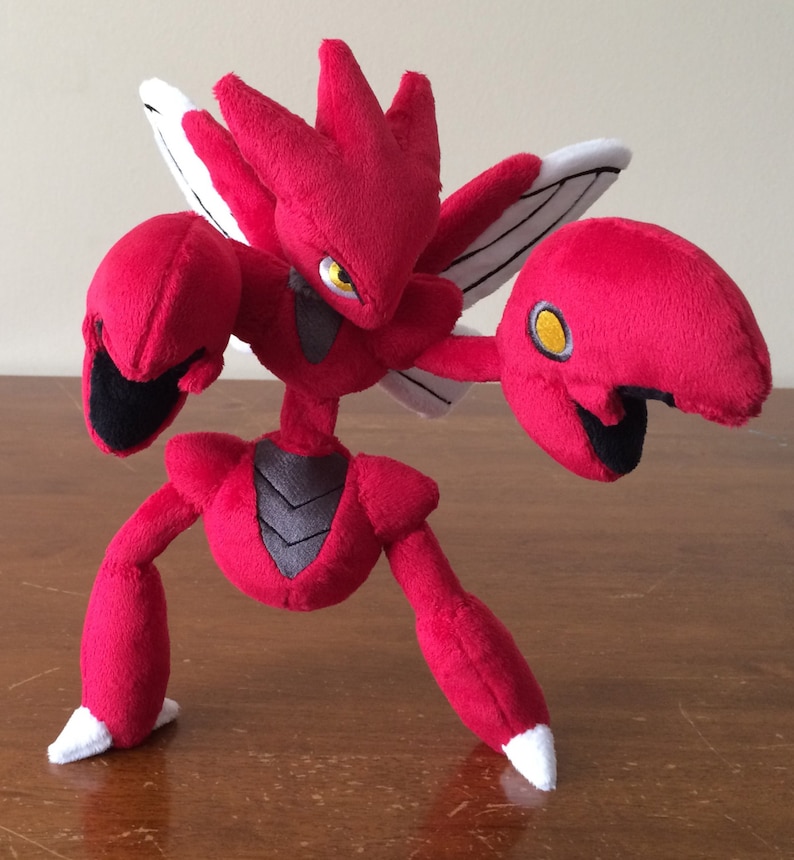 K&ouml;nnte beinhalten: Rotes und schwarzes Pl&uuml;schtier eines Pokemon-Charakters mit gro&szlig;en Krallen und Fl&uuml;geln. Das Spielzeug steht auf einer Holzoberfl&auml;che.