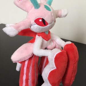 May include: Pink and red plush toy with a white face and green horns. The toy has a red eye and a white body with red stripes. The toy is holding its arms in front of it.