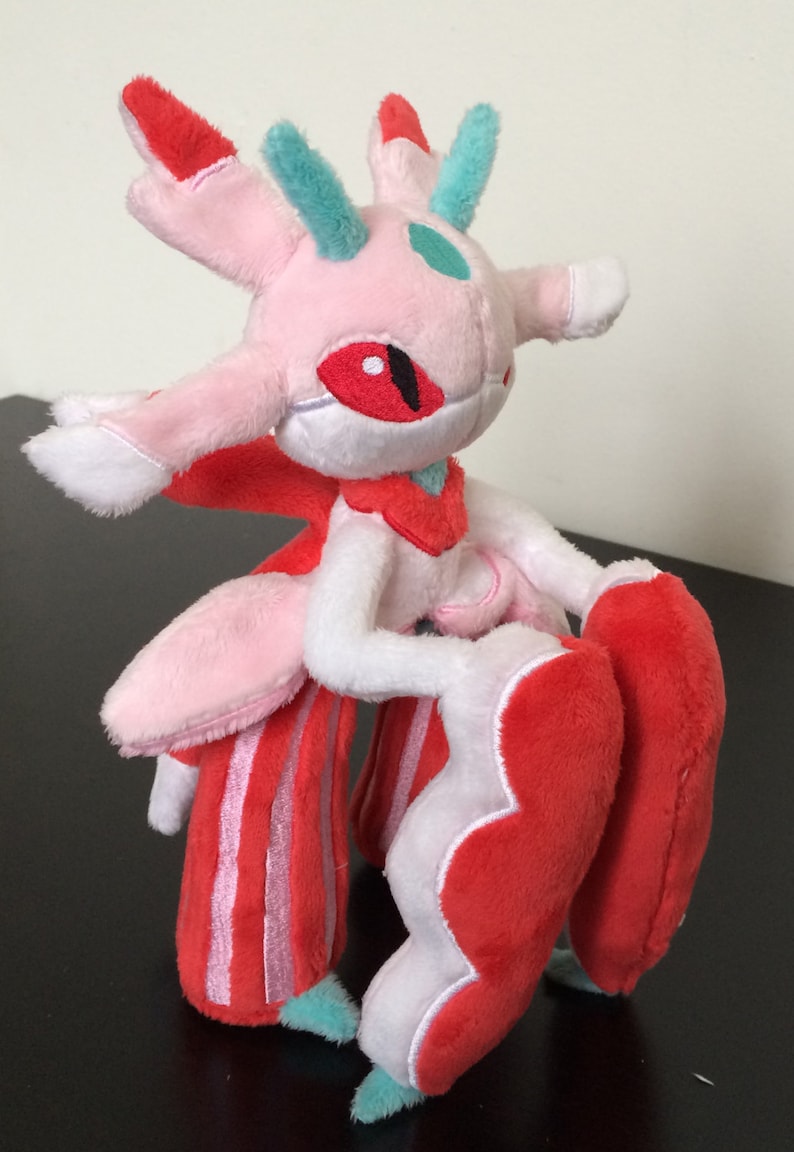 May include: Pink and red plush toy with a white face and green horns. The toy has a red eye and a white body with red stripes. The toy is holding its arms in front of it.