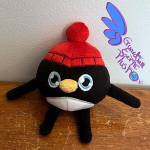 Puede incluir: Un juguete de peluche de ping&uuml;ino blanco y negro que lleva un gorro de punto rojo con un pomp&oacute;n blanco. El ping&uuml;ino tiene un pico amarillo y ojos azules.