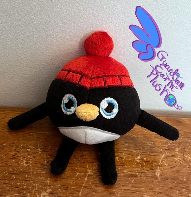 Puede incluir: Un juguete de peluche de ping&uuml;ino blanco y negro que lleva un gorro de punto rojo con un pomp&oacute;n blanco. El ping&uuml;ino tiene un pico amarillo y ojos azules.