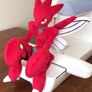 K&ouml;nnte beinhalten: Ein rotes und wei&szlig;es Pl&uuml;schtier eines Pokemon-Charakters mit Fl&uuml;geln. Das Spielzeug sitzt auf einer wei&szlig;en N&auml;hmaschine.