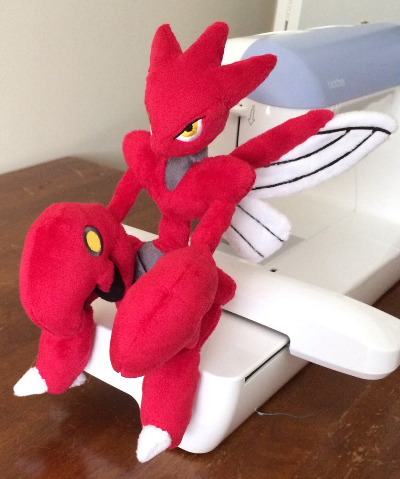 K&ouml;nnte beinhalten: Ein rotes und wei&szlig;es Pl&uuml;schtier eines Pokemon-Charakters mit Fl&uuml;geln. Das Spielzeug sitzt auf einer wei&szlig;en N&auml;hmaschine.