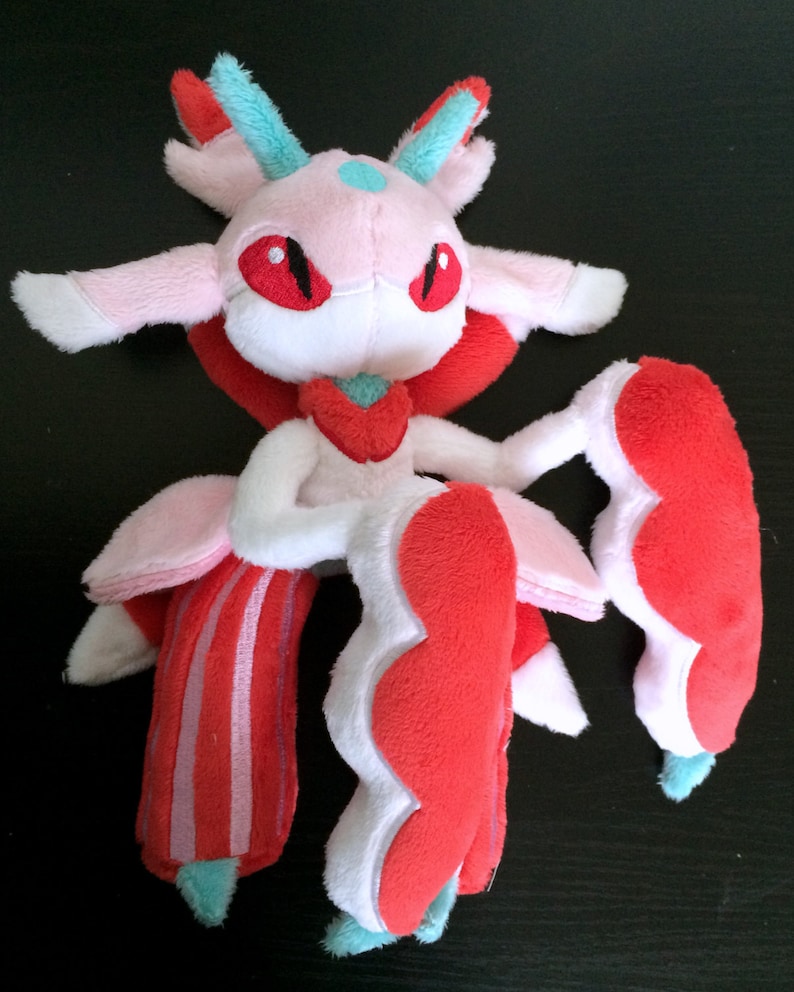 May include: A pink and red plush toy with a white face and red eyes. The toy has a long, flowing tail and a large, fluffy head.