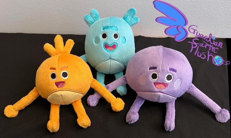 Puede incluir: Tres peluches, uno azul, uno naranja y uno morado, con caras y extremidades bordadas. Los juguetes sonr&iacute;en y tienen ojos grandes. El juguete azul tiene cuernos, el juguete naranja tiene una cabeza puntiaguda y el juguete morado tiene una cabeza redonda. El texto "Guardian Earth Plush" es visible en el fondo.
