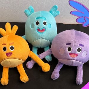 May include: Three plush toys, each with a different color and design. The first toy is orange with a smiling face and a spiky orange hair. The second toy is light blue with a smiling face and two antlers. The third toy is purple with a smiling face and a round body. The toys are all made by Guardian Earth Plush and are available to order.